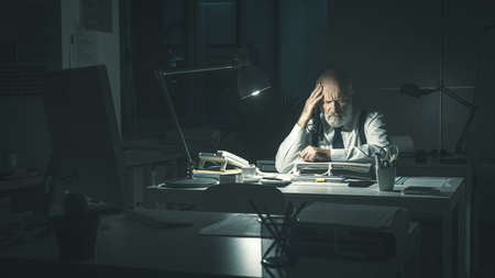 Stressed business executive sitting at desk and working overtime at night, he is having a bad headacheの写真素材