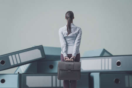 Businesswoman holding her briefcase and staring at a load of messy disorganized ring binders, back viewの写真素材