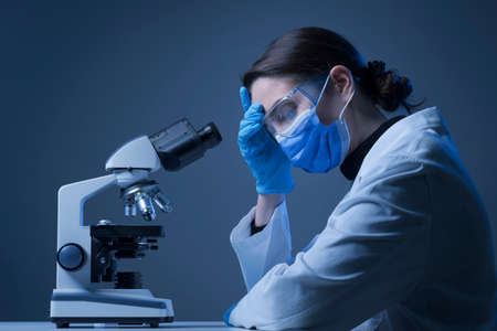 Pensive disappointed researcher with face mask in the lab, she is using a microscope: difficulties and challenges of medical researchの写真素材