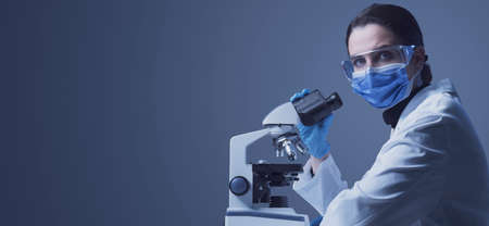 Researcher wearing a surgical mask and using a microscope, she is looking at camera: medical...