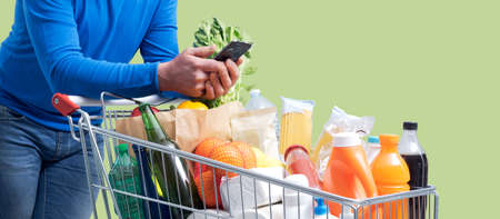 Man pushing a shopping cart and using a smartphone, grocery shopping and technology concept