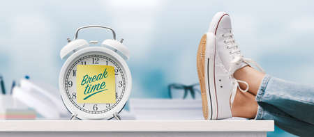 Woman relaxing with feet up in the office and alarm clock with sticky note, it's break time