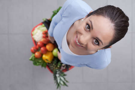 Young woman holding a shopping basket full of fresh vegetables and fruits, she is smiling at camera, top viewの写真素材