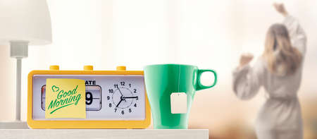 Alarm clock with good morning message and tea cup on the bedside table, a woman is stretching in the backgroundの写真素材