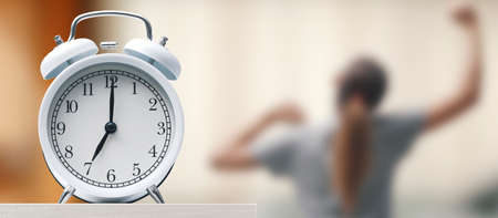 Alarm clock on the bedside table, a woman is getting up and stretching