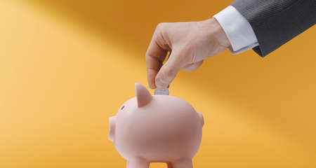 Corporate businessman inserting a coin in a piggy bank: saving money, deposits and investments conceptの写真素材