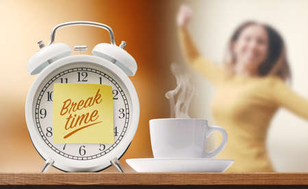 Happy young woman having a coffee break, alarm clock with sticky note and coffee cup in the foregroundの写真素材
