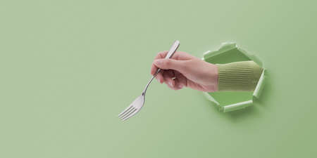 Female hand holding a fork and coming out of a hole in paper, eating and restaurants concept