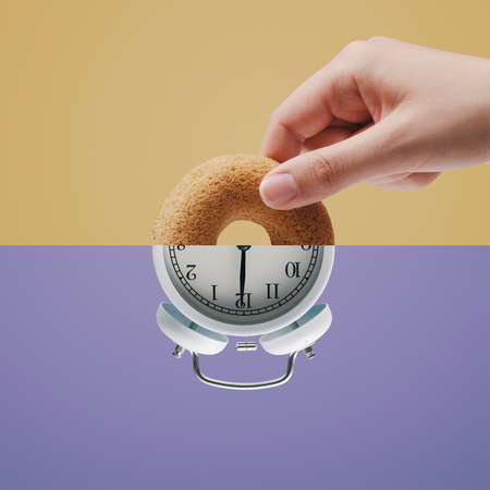 Half alarm clock and hand holding a donut shaped cookie, it's snack time, copy spaceの写真素材