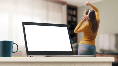 Laptop with blank screen and young woman dancing at home, she is wearing wireless headphones and...