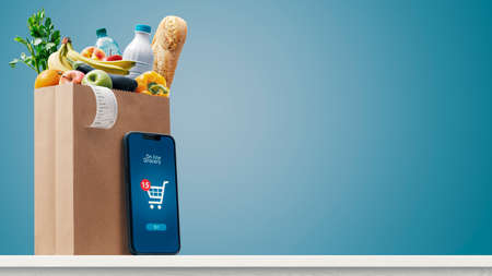 Grocery shopping app and grocery bag full of goods: online grocery shopping and home delivery concept, copy spaceの写真素材