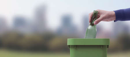 Woman putting an empty glass bottle in the trash bin, separate waste collection and recycling concept, natural landscape in the backgroundの写真素材