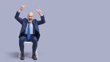 Cheerful businessman sitting on a chair and making V signs, he is celebrating his successの写真素材