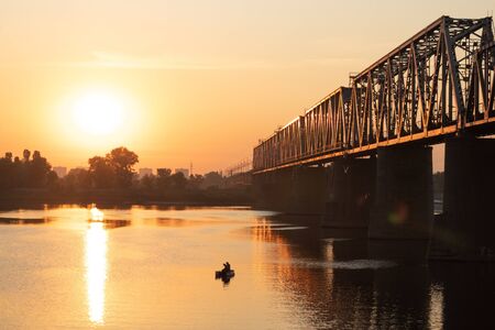 Sunrise. Railway bridge over a wide river. Dnieper River, the city of Kiev, Ukraine. Silhouette of a fisherman on a boat.の写真素材