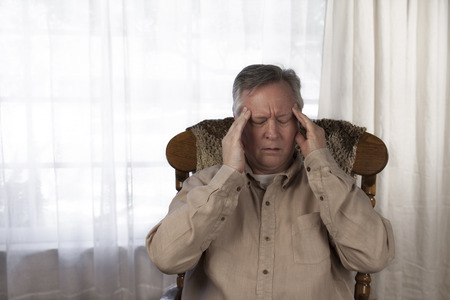 Elderly man holding his hands to his head as he suffers from a headache or migraine. Sitting at home in a rocking chairの写真素材
