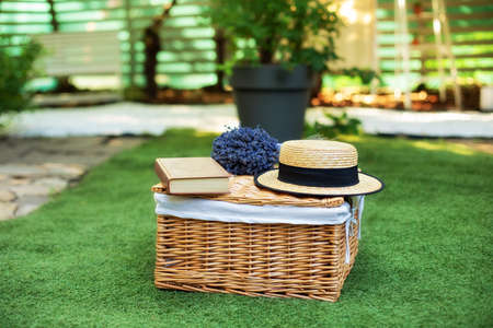 Wicker picnic basket with flowers on grass in garden. Weekend concept. Picnic basket with a book and a bouquet of lavender on the grass. Cozy Picnic lunch outdoors on the lawn on a sunny day in park.の写真素材