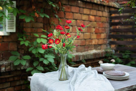 A bouquet of red poppies in a glass vase. Flowers in a vase at home. Interior Terrace, patio. Wooden table and plates for garden party or dinner. Table for lunch outside in garden in yard of house.の写真素材