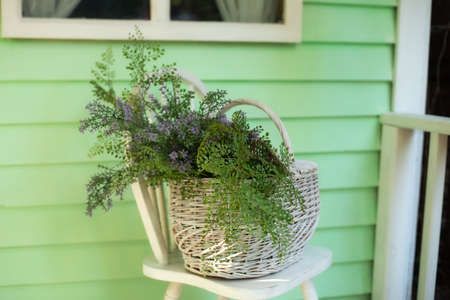Wicker basket with bouquet wild flowers on wooden chair near wall. Decoration in patio home. Autumn terrace with chair and basket with flowers. Stylish fall decor on front porch home. Space for textの写真素材