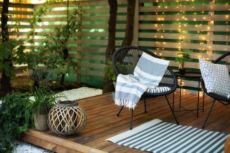 Exterior veranda of house with black Acapulco armchair and plants pots. Cozy space in patio or balcony with garland. Interior Wooden verande with garden furniture. Modern lounge outdoors in backyardの写真素材