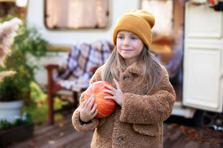 Happy Halloween. Smiling girl standing on the porch with a pumpkin in her hands. Trick-or-treat. Happy little girl playing near camping at RV home in fall garden. Child in cozy campsite fall backyard.の写真素材