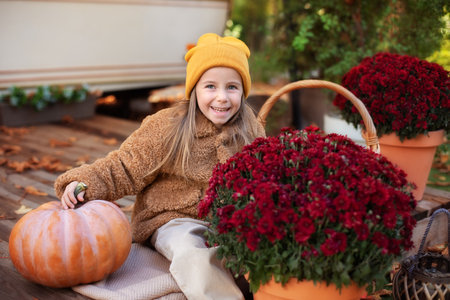 Happy little girl sitting on porch of house with chrysanthemums potted and pumpkins. home fall decoration for Halloween or Thanksgiving. Smilling Child in autumn garden with yellow pumpkins and flowerの写真素材