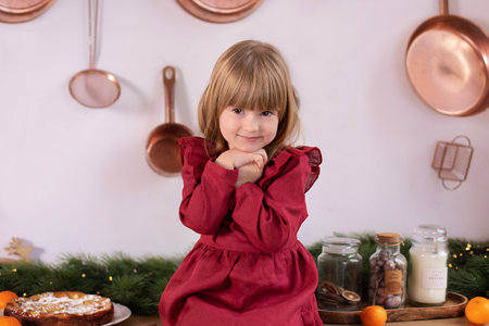Happy little girl in red dress on Christmas eve at home. Happy little girl sitting on tabletop on kitchen with christmas pie and tangerinesの写真素材