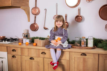 Smiling little girl sitting on the wooden table top on kitchen with christmas gifts and tangerines. Child plays in decorated kitchen for Xmas. Happy little girl in dress on Christmas eve at homeの写真素材