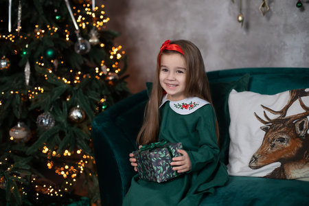 Smiling happy little girl in green dress by Christmas tree with glowing garland lights. Beautiful small girl in living room opens present on Xmas Eve.の写真素材