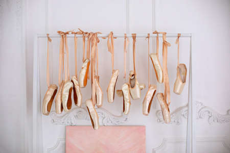 Many hanging ballet shoes on white wall background in studio. New pointe shoes with satin ribbons hanging on rank. Ballet shoes hang on bar in room. Concept of dance, ballet school, ballerinas clothesの写真素材