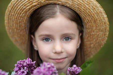 Smiling child girl with big eyes looking in camera. Closeup portrait of a romantic charming little girl in straw hat. Childhood concept. Cute portrait of a caucasian smiling girl with summer flowers.の写真素材