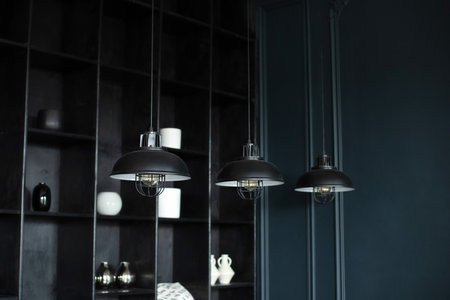 Three modern black ceiling lamps hang in room, close-up. loft room interior. Metal chandeliers in retro style on background wooden shelves with decor.の写真素材