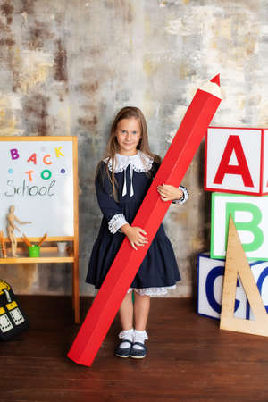 back to school! Happy smiling little girl in dress stands with large pencil in her hands at school. Schoolchild is studying in the classroom on the background of the school board.の写真素材