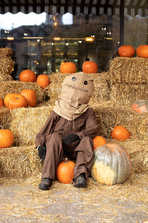 Scary creepy scarecrow with with a bag of straw for a head in overalls sit on stack of hay or straw with many orange pumpkins. Halloween decoration at home.の写真素材