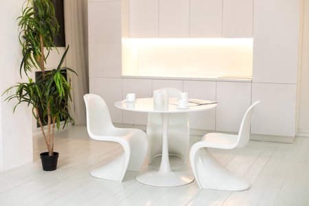 Modern Kitchen interior design with furniture. Stylish white dining room with large round table and designer chairs, cups coffee, large houseplant in pot. White plastic chairs pantone.の写真素材