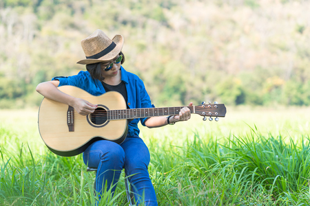 Young asian women short hair wear hat and sunglasses sit playing guitar in grass field countryside Thailandの写真素材