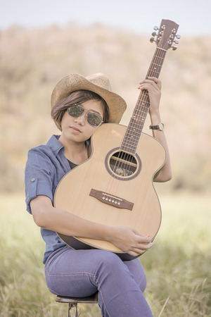 Young asian women short hair wear hat and sunglasses sit playing guitar in grass field countryside Thailandの写真素材