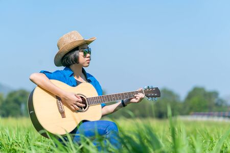 Young asian women short hair wear hat and sunglasses sit playing guitar in grass field countryside Thailandの写真素材