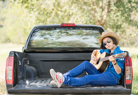 Young asian women short hair wear hat and sunglasses playing guitar ,sit on pickup truck in countryside Thailandの写真素材