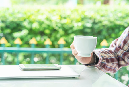 Male hands holding smart phone and cup of coffee on the table  outdoor with blurred green plant...