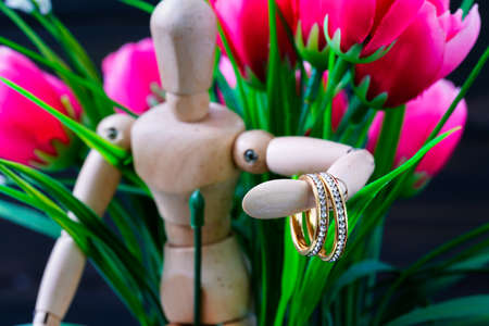 Wooden figure holding golden wedding ring resting with red rose flowerの写真素材