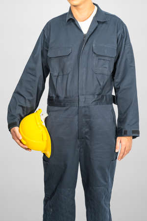 Worker standing in blue coverall holding yellow hardhat isolated on gray backgroundの写真素材