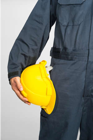 Worker standing in blue coverall holding yellow hardhat isolated on gray backgroundの写真素材