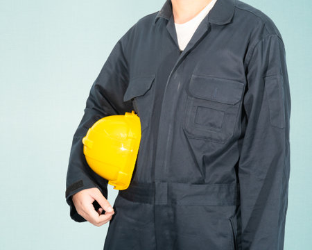 Worker standing in blue coverall holding yellow hardhat isolated on blue backgroundの写真素材