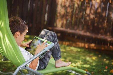 boy sitting with a tablet in the garden. the boy lies on a deck chair and plays on the tablet. child...
