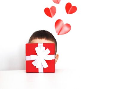 a young man holding a small present box with red hearts on a white background. child covers his face with a boxの写真素材