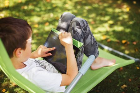 boy sitting with a tablet in the garden. the boy lies on a deck chair and plays on the tablet.の写真素材