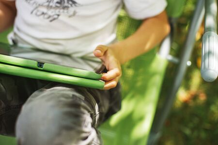 boy sitting with a tablet in the garden. the boy lies on a deck chair and plays on the tablet.の写真素材