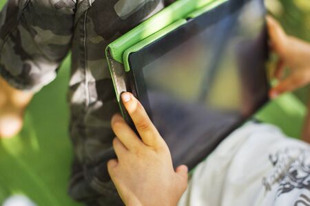 boy sitting with a tablet in the garden. the boy lies on a deck chair and plays on the tablet.の写真素材