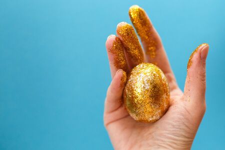 Easter golden egg in the hand on a blue background. Easter background. hand and egg in gold sparkles. happy Easterの写真素材