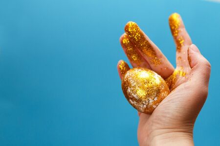 Easter golden egg in the hand on a blue background. Easter background. hand and egg in gold sparkles. happy Easterの写真素材